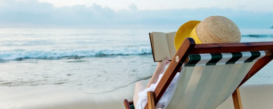 Person liest ein Buch auf einem Liegestuhl am Strand mit Blick auf das Meer, ideal für Urlaubslektüre
